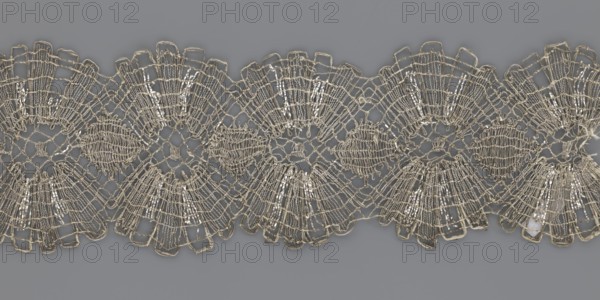 Lace fragment, metal threads, 18th century, Europe, lace fabric, width 8.2 cm, length 27.5 cm., A decorative length of lace with an intricate openwork pattern lies horizontally across a solid background; the lace shows a repeating scalloped design made of interlaced threads forming fanlike and circular motifs, with areas of dense stitching and more open netted sections, and small reflective elements woven into some parts. The visible colors are pale beige or off-white for the lace, metallic-silver highlights where the reflective elements catch light, and a muted medium gray for the background.
