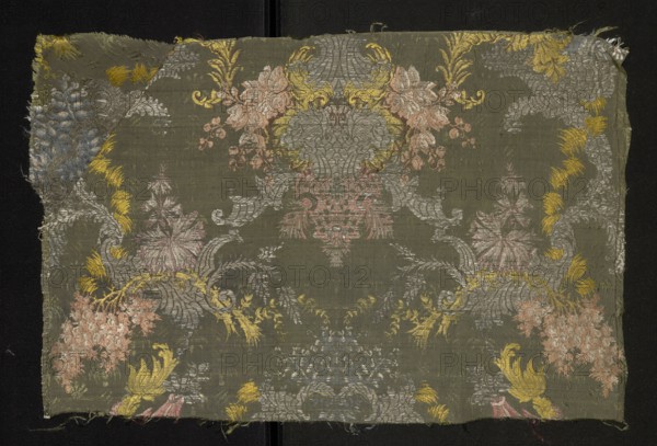 Fragment of fabric, fabrics, fragments of silk patterned fabric with floral patterns. Pattern with a symmetrical composition of fantasy-floral motifs: lilac branches entwined with silver leaves stylized into a Rococo ornament. Background: gros de Tours green, pattern brocaded with threads tied in a weft weave: lemon, celadon, light and dark pink, white, silver braided thread, brown. Piece width: 54.5 cm. Border width: 0.6 cm. 1st half 18th century, France; height 54.5 cm, width 36 cm., A rectangular fabric fragment with a muted olive-green background displays an ornate symmetrical floral and foliate pattern in a few colors including pale pink, soft gray, muted yellow, and a faded blue. The woven surface shows a central elaborate cartouche-like motif flanked by scrolling acanthus shapes and stylized blossoms, with clusters of rounded flowers and leafy sprays arranged across the cloth. The edges are slightly frayed and the weave texture is visible, with areas of metallic sheen in the gra...
