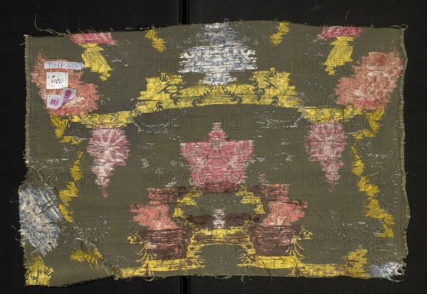 Fragment of fabric, silk, patterned with floral motifs. Pattern with a symmetrical composition of fantasy-floral motifs: lilac branches entwined with silver leaves stylized into a Rococo ornament. Background: gros de Tours green, pattern brocaded with threads tied in a weft weave: lemon, celadon, light and dark pink, white, silver braided thread, brown. 1st half 18th century, France. Piece height 54.5 cm, width 36 cm; piece width listed 54.5 cm; border width 0.6 cm., A rectangular piece of fabric with a dark olive-green background shows worn embroidered motifs in muted yellow, pink, pale blue, and brown. The fabric has a central pink ornamental shape flanked by symmetrical pink and pale blue decorative elements, connected by bands and scroll-like patterns in yellow. There are several frayed edges and loose threads around the perimeter, and areas where the embroidery appears abraded or missing, revealing the underlying weave. Small patches of pale blue and yellow appear near the corners...