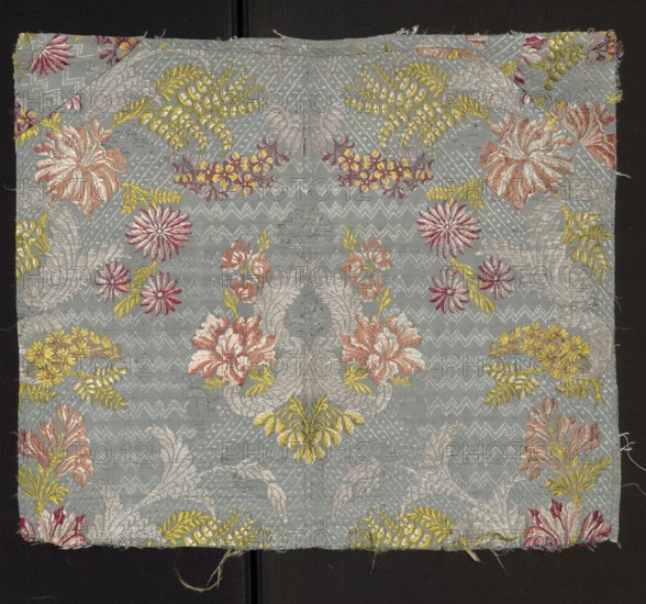 Fabric fragment: silk patterned with a symmetrical composition of branches of various stylized flowers surrounded by a geometric-floral overprint ornament. Blue background Gros de Tours, liseré; pattern brocaded with colorful threads in light green, yellow, white, blue, light and dark red, light brown, pink. Size of the repeat larger than the preserved fragment. Dimensions: length 38 cm report larger than fragment, preserved panel height 51.5 cm, width 44 cm; edge width 0.6 cm. 18th century, France., A square piece of woven fabric with a pale bluish-gray background covered in an ornate floral and foliate pattern in colors including pale pink, dusty rose, deep pink, yellow, gold, and silvery-white, with a repeating arrangement of stylized flowers, fernlike leaves, and curved acanthus shapes; the fabric shows surrounding raw edges and slight fraying, and it is placed against a dark background with a narrow vertical seam visible beneath the fabric.