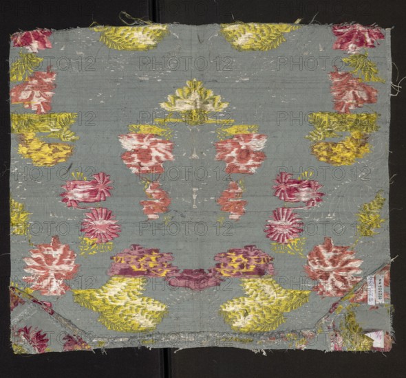 Fabric fragment. Silk brocaded with a symmetrical composition of branches of various stylized flowers surrounded by a geometric-floral overprint ornament. Background blue gros de Tours with liseré; pattern brocaded in light green, yellow, white, blue, light and dark red, light brown, and pink. Floral pattern. 18th century, France. Report size larger than preserved fragment. Preserved fragment dimensions: height 51.5 cm × width 44 cm; length of report approx. 38 cm; width of edge 0.6 cm., A square piece of fabric with a muted blue-gray background displaying a symmetrical floral pattern in shades of yellow, pink, red, white, and purple, with a few clustered flower shapes and leaf-like motifs repeated across the surface; the woven texture and some worn areas are visible, and frayed edges appear along the top and bottom with a folded corner at one side showing the fabric reverse; thin darker seams run vertically near the center, and scattered loose threads lie on and around the textile.