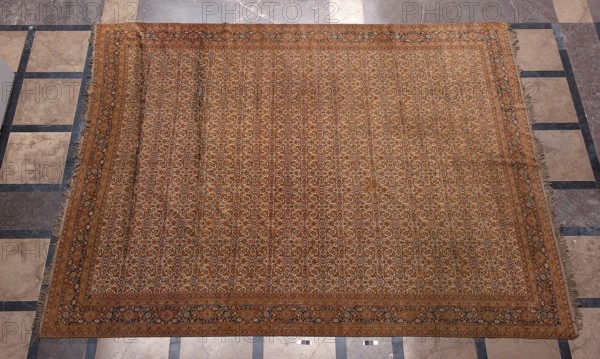 Carpet, Knotted decorative woven fabric carpet, 19th century, Iran. In the central field on a light cream background is a fine net ornament Herat säck composed of lanceolate leaves, rosettes, palmettes, stylized flowers, and rhombuses. Five-band border with floral motifs, the widest central band on a navy-blue background. Colors: brick red, red, green, blue, navy blue, and white. Height 520 cm; width 395 cm., A large rectangular patterned rug lies flat on a tiled floor, showing a dense repeating floral and geometric motif across its field with a decorative border and fringed edges. The rug's colors include shades of brown, tan, beige, rust, and muted orange, with accents of dark blue and black in the border and pattern. The surrounding floor tiles are a mix of light beige, gray, and darker gray tones arranged in a grid with darker grout lines. The rug shows slight ripples and variations in texture and the fringe appears slightly uneven along the sides.