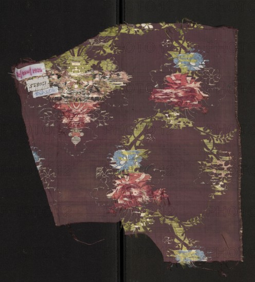 Fabric fragment, fragments of silk fabrics, patterned fabrics, floral patterns. Silk with a pattern of a symmetrical composition of bouquets and sprigs of multicolored stylized flowers. Gros de Tours background, liseré; pattern brocaded with colorful threads: green, white, blue, light and dark red, pink; silver threads thread with braid tied in rows. Report dimensions larger than the preserved fragment. Edge width: 0.3 cm. 2nd half of the 18th century, France. Height 34 cm, width 38 cm., A torn piece of woven fabric with a muted purple background featuring a repeating floral pattern in shades of red, pink, blue, yellow, green, white, and pale beige; the flowers appear in vertical garlands connected by green leafy vines and pale decorative motifs, with some areas showing frayed threads and worn patches where the weave is disturbed; the fragment rests against a dark black background and has irregular edges with loose fibers visible along the borders.