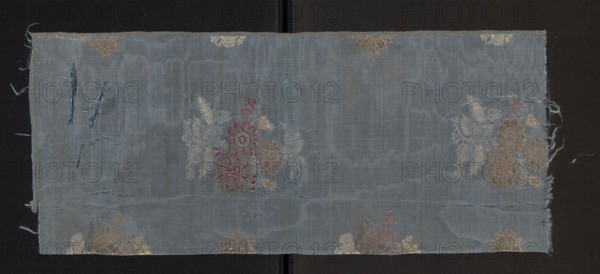 Fabric fragment, 17th century, Italy. Brocaded and morayon grosgrain silk. Weft grosgrain construction with blue silk warp and weft; pattern brocaded with silk threads held with a weft weave in dark pink, beige, salmon, white, and a gold-plated thread braided on a white silk core moriated. Design: loosely scattered smaller and larger floral sprigs on a blue background; stems and leaves in white with petals outlined in red and salmon-pink and filled with gold or left plain; probably larger bushes in two alternating-row types with smaller elements between. Motif size reported 14 x 10.5 cm; preserved fragment dimensions height 57 cm, width 24 cm; selvedge 0.4 cm wide blue and beige silk threads. Weaving edge: 0.4 cm wide silk with blue and beige stripes., A rectangular piece of worn woven fabric lies flat against a dark background, showing a muted blue-gray ground with visible vertical and horizontal weave texture and areas of fraying along the edges; several floral motifs in faded gold, ...