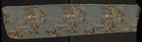 Fabric fragment, silk fabric, patterned, preserved selvedge, Taffeta background in blue-green, brocaded with colorful threads tied in a twill weave. Brocade colors: light and dark greens, browns, pale pinks, dark cherry, blue, gray, silver, olive green, lemon green, reddish brown. Floral motifs and fragments of garden architecture. Dimensions: length 26 x ?, edge width 0.5 cm, height 111 cm, width 21 cm. 1730s, France., A narrow strip of woven fabric with a muted blue-gray background displaying a repeating decorative scene in soft, faded colors; the design includes a group of stylized floral and leafy forms in shades of yellow, pink, red, green, cream, and brown, along with architectural elements resembling a low wall or terrace worked in pale gray and brown tones, and rounded tree-like shapes in pale green and beige. The fabric shows frayed edges and small loose threads along the top and bottom, with areas of slight discoloration and faint surface marks in darker gray and brown. The p...