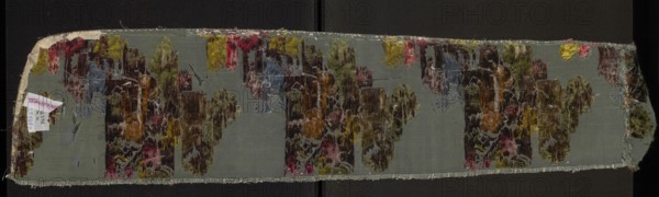 Fabric fragment, silk taffeta background in blue-green, brocaded with colorful threads in a twill weave. Brocade colors: light and dark greens, browns, pale pinks, dark cherry, blue, gray, silver, olive green, lemon green, reddish brown. Patterned with floral motifs and fragments of garden architecture; preserved selvedge. French, 1730s. Dimensions: height 111 cm, width 21 cm; fragment length 26 cm; edge width 0.5 cm., A long rectangular fabric strip laid flat on a dark background with frayed edges and irregular ends, showing a patterned pile on a smooth base; the base fabric appears gray-green, and the pile contains a mix of colors including olive green, deep brown, mustard yellow, muted pink, faded red, blue-gray, and touches of beige and white, arranged in several repeated abstract floral or cloud-like motifs; the pile shows worn and tufted areas with visible threads and some loose fibers, and a small folded label is attached near one short edge.