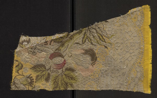 Fabric fragment, fragments of silk patterned fabric, 18th-century silk, fabric with preserved selvedge. Gros de Tours background — yellow brocaded with colorful threads: light and dark pink, light and dark salmon, light and dark green, dark salmon, cherry red, silver braided thread, silver lamellar. Threads tied with a twill weave. Pattern: fancy fruits against a silver lace pattern. Hem width: 0.9 cm. 18th century, France. Height 39 cm, width 24 cm., A worn rectangular textile fragment with frayed edges rests against a dark background; the fabric shows a decorative woven pattern with floral and foliate motifs and a scalloped border area. The visible colors include yellow, gold, olive green, brown, beige, pale pink, red, and silver-gray. There are large stylized leaves and petal shapes, textured diagonal weaving, and areas with metallic-looking thread that create a shimmering effect. The right edge has a brighter yellow selvedge, while other edges show loose threads and slight discolor...
