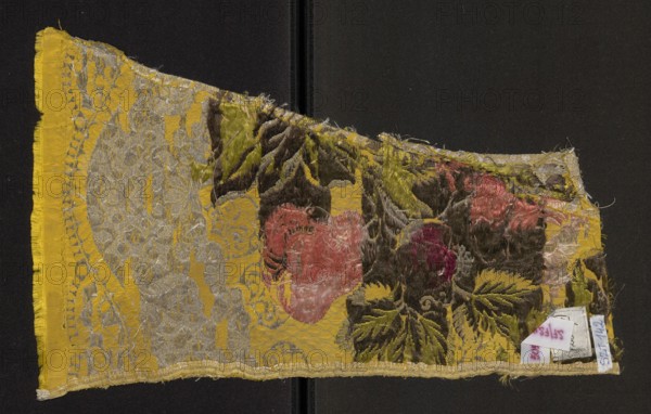 Fabric fragment, fragments of silk patterned fabric, 18th-century silk, fabric with preserved selvedge. Gros de Tours background — yellow brocaded with colorful threads: light and dark pink, light and dark salmon, light and dark green, dark salmon, cherry red, silver braided thread, silver lamellar. Threads tied with a twill weave. Pattern: fancy fruits against a silver lace pattern. Hem width: 0.9 cm. 18th century, France. Height 39 cm, width 24 cm., A worn textile fragment lies against a dark background, showing a torn, irregular triangular shape with frayed edges. The fabric displays a bright yellow ground with a patterned area of metallic-looking threads forming an ornate, lace-like design. Over parts of the yellow are raised, velvety patches in olive green, dark brown, and deep maroon, with a few areas of faded pink and muted red forming floral-like shapes. A narrow yellow border runs along one long edge, and parts of the pile appear flattened or worn, revealing lighter and darker...