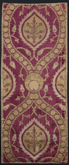 Fabric piece, Repossession, Turkish fabric, 16th century. Red sheared velvet ground with cream and silver patterns in satin and weft weaves. Large repeat of pointed ovals formed by wide ribbons fastened with a peacock's eyes motif; ribbons filled with single circles. Inside ovals: palmettes with a central bouquet of flowers. Surrounding the ovals: winding stems of small flowers. Height 154 cm, width 63 cm., The image shows a vertical textile with a repeating ornamental pattern in deep magenta and muted gold tones on a dark background border. The design features a series of large teardrop-shaped medallions with pointed leafy outlines containing floral motifs, connected by curved bands decorated with a row of circular elements. Stylized leaf and bud shapes extend from the bands and fill the surrounding field. The textile surface shows areas of wear and slight discoloration where the magenta and gold threads vary in intensity, and the edges appear slightly frayed.
