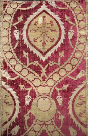 Fabric piece, fabrics, Turkish, velvets, net-composition patterns, large repeat of pointed ovals formed by wide ribbons fastened with a peacock's eyes motif. Ribbons filled with single circles; inside the ovals are palmettes with a central bouquet of flowers. Around the ovals are winding stems of small flowers. Red velvet background with cream and silver patterns. Satin and weft weaves; sheared velvet. 16th century, Turkey. Dimensions: height 154 cm, width 63 cm., The image shows a patterned textile with a dominant deep red background and large areas of muted gold forming ornamental shapes. The design includes a central medallion shape with jagged edges, floral and vine motifs, circular linked elements, and teardrop or bud-like shapes. The gold patterning contrasts with the worn red velvet-like background, and the textile surface shows areas of wear and texture variation. The overall composition is symmetrical with repeating decorative motifs in gold against the red field.