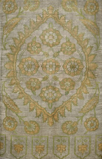 Fabric slab, fragments of a larger textile object, silk fabric. In the center, a large palmette covering the full width of the slab, edged with small serrated leaves; inside, symmetrically arranged flat flowers rosettes. In the corners, flower sprigs. On one narrower edge, a row of niches each with a central flower. Ornament colors: silver background, gold, and green. 16th century, Turkey. Height 103 cm, width 68 cm., The image shows a textile with an ornate floral pattern in shades of green and gold on a muted gray-beige background; a central medallion of stylized flowers and leaves is surrounded by a garland-like border, with additional floral motifs and elongated leaf shapes radiating outward, and a row of shield-like panel motifs along the lower edge, all rendered with a slightly raised, textured appearance.