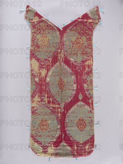 Back of the chasuble, fabrics, metal threads, liturgical vestments, Turkish fabrics, fragments of silk fabrics, patterned fabrics, flowers, leaves, plant ornaments, church paraments, sacred art, churches and chapels furnishings. The back of the chasuble is sewn from two fragments of fabric with motifs of large palmettes placed on a plain red background. The palmettes with serrated edges are decorated inside with floral sprigs and fruit spreading symmetrically and enclosing a smaller palmette with arabesque patterns in the center. Patterns in cream, blue, and gold. Three pieces of fabric sewn into one. Regular plant ornament, unfolding vertically on a red background. Colors: gold, white and sapphire. 17th century, Turkey, liturgical vestment, length 118 cm, width 49 cm back., A long, narrow textile panel with a V-shaped top and rounded bottom, displayed flat against a pale background, showing a repeating ornate medallion pattern in a dominant deep red with accents of light blue and pale...