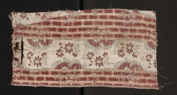 Fabric fragment, silk, patterned with floral motifs. Main motif: a wide, winding white satin ribbon projecting flower sprigs with striped petals. Ribbon background shows alternating dark and light salmon-pink horizontal stripes; striped ground includes small white flowers, larger flower sprigs, and regularly spaced vertical pink stripes. Ground is satin with warp-faced fabric; ornament executed in plain weave. Colors: background in four shades of pink, transverse stripes of twill and pink satin, white satin ribbon along the length. Warps in white and pink; wefts bound with a twill weave in three shades of salmon pink and white. Dimensions of fragment: 18 x 17.5 cm; edge width: 0.5 cm. Date: second half of the 18th century; probable origin: Sweden. Overall dimensions noted: height 43 cm, width 22 cm., A rectangular piece of worn fabric laid flat against a dark background shows a repeating floral and stripe pattern; the fabric has a pale base color with floral motifs in a muted red and l...