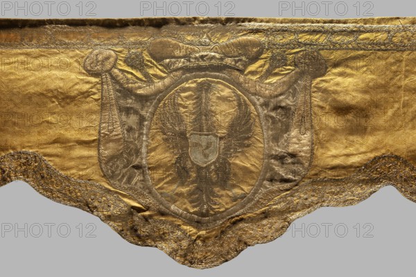 Drum apron or cauldron of the Polish cavalry of Prince Michal Radziwill, Polish Lithuanian Hetman 1668–1680?? Initials and coats of arms appliqued and embroidered in shields on yellow damask; features Traby coat of arms, Radziwill family monograms; ca. 1750, Poland; fabrics; height 43 cm min. 27.5 cm, width 205 cm., A scalloped textile panel with a metallic sheen occupies the image, predominantly gold in color with areas of darker, muted brownish-gold forming decorative patterns; the central area shows an oval medallion in a slightly darker tone containing a stylized double-headed bird motif rendered in deep brown-gold, and a small shield-like shape at the bird’s center in a paler, slightly bluish-gray tone; surrounding the medallion are draped, banner-like shapes in a darker brown-gold with subtle floral or brocade textures, and the top edge features a narrow decorative band in darker gold; the lower scalloped border is richly embroidered or embellished in darker brown-gold and gold t...