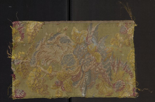 Fabric fragment, fabrics, fragments of silk fabrics, patterned fabrics, three-dimensional patterns, plant patterns. Background: green grodetur cannetillé; brocade pattern: yellow, olive, light and dark pink, green, salmon, white, brown, metal thread with silver braiding. In the Louis XVI style, fruit and flowers on a green background, 18th century, France; fabrics; height 44 cm, width 27 cm., A rectangular textile swatch with a woven floral and foliate design sits against a dark background; the fabric shows a palette of olive green, mustard yellow, muted gold, dusty blue, pale beige, soft pink, and touches of faded red, with areas of worn fraying at the edges and corners, a folded triangular patch at one corner, and a central cluster of stylized flowers and leaves rendered in layered threads that create textured, raised motifs across the surface.
