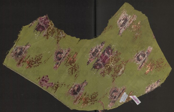 Fabric fragment, silk, unknown origin, 18th-century silk fragment. Pattern: symmetrical composition of flower bouquets and a lace ribbon. Ground: grass-green background grosgrain weave with a delicate, fanciful drawing of clouds—leaves achieved by releasing weft threads. Embroidery/broaching colors: blue, olive, white, pale pinks and reds, cherry red, cream, salmon, black. Measurements: reported 29.5 x ?; selvedge width 0.6 cm; overall height 32 cm, width 77 cm. Date: second half of the 18th century; possible Russia., A worn piece of fabric with an irregular curved shape lies against a dark background, predominantly olive green with patches of faded pink, burgundy, brown, beige, and cream forming floral and abstract motifs; the fabric shows frayed edges and loose threads, areas of abrasion and thinness, and several clusters of brushy, textured color where the weave is disturbed, while a small folded cloth tag of pale color is tucked near one edge.