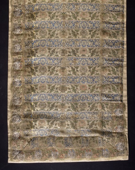 Fragment of a belt with a continuous pattern of alternating silhouetted tendrils with an iris motif and colorful tendrils with saxophone leaves and symmetrical palmettes. Border of floral tendrils and medallions with portraits of men in turbans. Background of the continuous belts: navy blue and gold-lipped. Background of the border belt: gold-lipped; background in the cartouche with a male bust - silver-white. The silhouetted motifs in the shelves are similar to other known Persian belt patterns. The warp is made of salmon-colored silk yarn; the weft is gold metal thread wrapped around a silk thread. Preserved only in fragments; part of the hem was cut from the body and sewn along the length of what was likely a severed head. Early 17th century, Iran; clothing accessory; height 115 cm, width 32 cm., A rectangular woven textile laid flat against a dark background, showing horizontal patterned bands with floral and foliate motifs in metallic gold and muted beige, with accents of blue, gr...