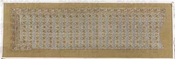 Fragment of a belt with a continuous pattern of alternating silhouetted tendrils with an iris motif and colorful tendrils with saxophone leaves and symmetrical palmettes. Border of floral tendrils and medallions with portraits of men in turbans. Background of the continuous belts: navy blue and gold-lipped. Background of the border belt: gold-lipped; background in the cartouche with a male bust – silver-white. The silhouetted motifs in the shelves are similar to patterns in other contemporary belts. The warp is made of salmon-colored silk yarn; the weft is gold metal thread wrapped around a silk thread. Preserved only in fragments; part of the hem was cut from the body and sewn along the length of what was likely a severed head. First half of the 17th century, Iran, clothing accessory; height 115 cm, width 32 cm., A rectangular textile fragment with a decorative striped pattern laid on a plain tan backing; the fragment shows alternating vertical bands of floral motifs in muted blue, gr...