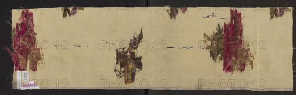 Fragment of fabric, patterned silk fabric with preserved selvedge, floral patterns, 18th-century French silk. Description: white cream background - gros de Tours liseré, brocaded with threads in olive green, dark green, dark pink, pale red, heather. Inscription/label: Gros de tours liseré, 1 lat de lancé, broché a liagé repris. Dimensions: height 81 cm, width 23 cm; fragment 59 x ?; selvedge width 0.6 cm., A long rectangular piece of pale beige fabric with a textured weave lies against a dark background; there are several torn and frayed floral patches in shades of deep red, burgundy, brown, olive green, and muted yellow scattered across the surface, including a tall vertical distressed patch near one short edge, a similar vertical patch near the opposite short edge, and a smaller vertical cluster near the center, with a few smaller fragmented floral fragments near the top edge; thin horizontal dark navy lines and small dark smudges interrupt the beige ground in places, and loose fiber...