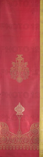 Tapestry, Turkish, late 17th century. Fragment trimmed on one side with an olive-colored silk border. At the top a stylized plant ornament forms a mihrab niche; a lamp hangs from the top of the niche. Below, in the center, a vase with a stylized bouquet of flowers in a symmetrical arrangement. Plain red plain-weave background; pattern brocaded with braided silver metal thread. Silk fabrics; height 280 cm, width 76 cm., A vertical piece of fabric with a predominant deep pink or magenta field and a narrow gold vertical border along the right edge, featuring ornate gold woven motifs: a large floral-medallion motif near the upper center, a smaller vase-like motif lower down connected by a decorative stem, and an ornate scalloped band with circular floral designs and a fringe-like patterned border running near the bottom; the fabric shows slight variations in texture and subtle darker and lighter areas, and the visible colors are deep pink or magenta and gold.