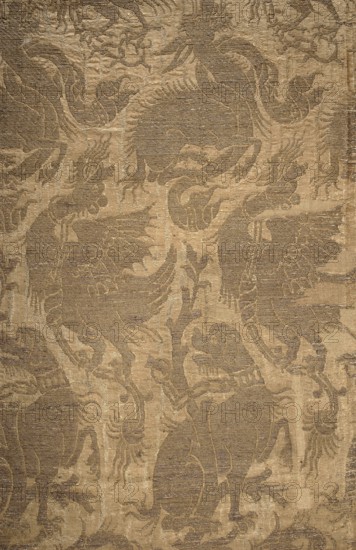 Fabric fragment, patterned silk brocaded with gold Cypriot thread on pale pink ground. The brocaded design forms a net of silhouettes of fantastic animals entwined with chains and ribbons. Among five animal types are identifiable dogs, winged dragons, and ambiguous beasts with the head of a donkey and the body of a snake. Motifs draw on ancient Far Eastern decorative art and medieval bestiaries; animal symbolism derives from ancient/tribal sources and zoological bestiaries physiologi and intersects with Christian symbolism. 14th century, Italy. Height 29 cm, width 84 cm., A textured fabric surface with an overall pattern of stylized birdlike figures and flowing ornamental shapes, rendered in shades of tan, beige, brown, and gold, showing raised areas and recessed stripes that create a subtle contrast between matte and slightly shiny threads, with repeating wing, tail, and crest motifs intertwined across the surface.