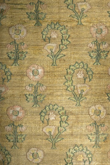 Tapestry, sewn from two fabrics. Rectangular central field of silk ikat fabric, interwoven with silver lamellar, with a check motif. Pattern on the shorter sides enclosed by strips of elongated triangles with an ikat effect. In every other field of the check, in eight vertical rows, gold lamellar brocade in a geometric square ornament. Border made of semi-silk fabric with brocade motifs of flower bushes in two types, in alternating rows a twig with a cotton flower and with angles and an arched shoot. Colors: central field light brown, white, yellow, turquoise blue; yellow-gold border with a white-pink-green-celadon pattern. 18th century, Iran. Materials: fabrics; height 245 cm, width 116 cm., The image shows a patterned textile with a gold background and a repeating floral motif. A group of stylized flowers and leaves are woven in soft pink, pale yellow, and muted green with outlines and stems in a darker greenish-blue. The fabric surface has a horizontal ribbed texture and the woven f...