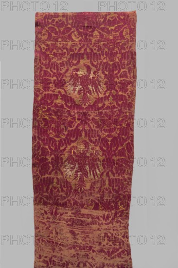 Fragment of fabric, 16th century, Italy. Brocade fragment sewn from several smaller pieces, woven with a net composition of interwoven ribbons forming reserves that include coats of arms Jagiellonian Eagle and bouquets of flowers in vases. Fabrics with preserved report, preserved ply width, and preserved selvedge. Predominant colors: carmine red, yellow, and white; background originally ochre . Height 234 cm, width 56 cm., A rectangular textile with a patterned surface in shades of deep red and gold is shown against a neutral gray background. The textile displays a repeating ornate motif with areas of metallic gold sheen interwoven with the red, creating a contrast between the two colors. The fabric appears textured with variations in color intensity, including patches where the gold is more prominent and areas where the red dominates. The overall piece is long and narrow with slightly uneven edges and visible folds and creases.