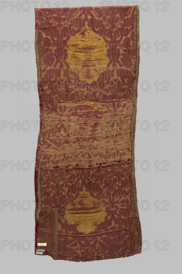 Fragment of a ply of fabric with a net composition, 16th century, Italy. In the reserves created from interwoven ribbons are the coats of arms: Jagiellonian Eagle and bouquets of flowers in vases. The fabric is sewn from several smaller fragments. Predominant colors: carmine red, yellow, and white; the background was originally ochre . Brocades; flowers; flowers in vases; patterned fabric with preserved report, preserved width of the ply, and preserved selvedge. Height 234 cm, width 56 cm., A long narrow textile panel with a worn patterned surface showing a repeating ornamental motif, predominantly maroon and golden yellow with traces of green along the narrow edges; the fabric appears aged and frayed in places with areas where the golden yellow threads are more exposed and raised, creating irregular textured patches; the pattern alternates between denser maroon ground and lighter golden designs that form central medallike shapes and surrounding floral or foliate impressions; the edges...