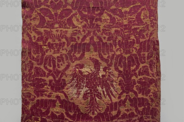 Fragment of fabric, 16th century, Italy. Brocade fragment sewn from several smaller pieces, a ply of fabric with a net composition. In reserves formed by interwoven ribbons are coats of arms Jagiellonian Eagle and bouquets of flowers in vases. Preserved features: patterned fabric with report, preserved ply width and selvedge. Predominant colors: carmine red, yellow, and white; background originally ochre . Height 234 cm, width 56 cm., A rectangular piece of fabric with a repeating ornamental pattern in deep red and muted gold tones, showing a central motif resembling a stylized winged shape formed by the red against the gold background; the textile has an overall textured, worn surface with irregular edges and areas where the gold appears more pronounced or faded, and the pattern consists of flowing, curvilinear shapes and abstract forms distributed across the fabric.
