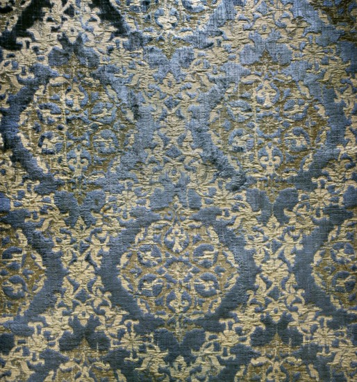 A fabric fragment: a diagonal net of plant and floral tendrils against a blue velvet background. In the fields of the net, in the shape of pointed ovals, are rosettes composed of stylized plant forms. Colors: blue background; ornament sand-colored with gold thread. Materials: velvet and silk; fabrics with preserved selvedge and preserved ply width. Dimensions: fragment 30 x 44 cm; ply width 63 cm; selvedge width 1.3 cm. Date: 15th century, Spain., A close-up of a textured fabric or rug showing an ornate repeating pattern with rounded medallion shapes and intricate floral and scroll-like motifs; the surface has a raised, plush pile contrasting with flatter woven areas, creating visible shadows and highlights. The dominant colors visible are blue and gold with areas where the blue appears darker and the gold ranges from pale to deeper tones. The pattern repeats across the image with symmetrical arrangements and varying pile heights that give a slightly distressed or worn appearance in pl...