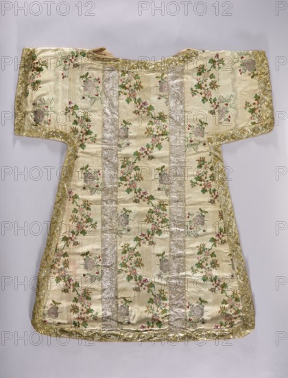 Dalmatic, origin, 18th century, France; liturgical vestment fabric decorated with flowers, leaves, and plant ornaments; used as church parament for churches and chapels; length 101.5 cm, width 92 cm back., A flat garment laid out against a light background, showing a T-shaped silhouette with short wide sleeves and a slightly flared lower section; the main fabric is a pale cream color with an overall pattern of floral clusters in shades of green, pink, and muted red, accompanied by greyish floral or urn-like motifs; there are several vertical bands of a shimmering silver fabric running down the front; the edges and sleeve hems are finished with a metallic gold trim that has a textured, embroidered appearance; the fabric shows subtle horizontal ribbing and slight irregularities in sheen where the light hits it.