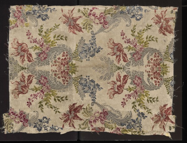 Fabric fragment, patterned silk fabrics, 18th-century France. Symmetrical decoration with axis through center of panel; fanciful floral sprigs entwined with silver and S-shaped leafy forms focused on axis of symmetry. Cream background woven with a grosgrain weave with delicate floated subreport flowers. Colors: white, pale pink, orange rose, rusty red, celadon yellow, grass green, dark green, pale blue, blue. Border width: 0.5 cm. Repeat: 39.5 x approx. 53 cm. Overall height 69 cm, width 53 cm. fragments of silk fabrics., A rectangular piece of woven fabric with frayed edges laid on a dark background, featuring an ornate repeating floral and foliate pattern in colors including pale beige background, muted blue, dusty rose, soft red, olive green, and gray; the design shows stylized blossoms, leafy sprigs, and curved paisley-like forms arranged symmetrically across the cloth, with several clusters of small rounded flowers, larger petaled flowers with flowing leaves, and scrolling vine el...