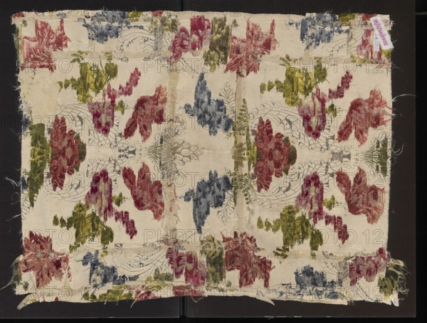 Fabric fragment, silk fabric fragment, patterned with fanciful floral sprigs entwined with silver and S-shaped leafy forms arranged symmetrically along a central axis. Colors on a cream grosgrain background with delicate floated subreport flowers: white, pale pink, orange rose, rusty red, celadon yellow, grass green, dark green, pale blue, blue. Border width 0.5 cm. Repeat: 39.5 x approx. 53 cm. 18th century, France. Overall dimensions: height 69 cm, width 53 cm., A worn rectangular piece of fabric with frayed edges lies against a dark background, showing a repeated floral and scroll motif in a faded palette; the base of the fabric is a creamy off-white with a few areas of discoloration, and the decorative sections are rendered in muted red, olive green, and dusky blue threads with visible brushing and pile texture, arranged symmetrically across the surface; outlines and ornamental curves are visible in a faint grayish tone, and there are several seams and patched areas where pieces of...
