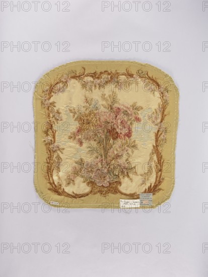 Two tapestry armchair backrest upholsteries and one sofa upholstery, varietal, flowers, floral ornaments, furniture upholstery, interior furnishings, decorative fabrics, tapestries; a, b armchair upholstery with a bouquet of flowers in the center, surrounded by a frame of acanthus flowers and leaves; c sofa upholstery with a spreading bouquet of flowers surrounded by a frame with acanthus flower and leaf motifs. Colors: sandy background, olive, pink, red, ochre, blue, and green ornaments. 18th/19th century, France; fabrics; height 64 cm, width 62 cm., A square textile with rounded corners is centered on a pale background; the textile is primarily yellow and beige with frayed edges and a decorative woven floral scene in the center showing a bouquet of flowers in shades of pink, red, brown, green, and muted blue, surrounded by an ornate border in brown and gold tones with additional small floral motifs, and a few small rectangular tags attached near the lower edge.