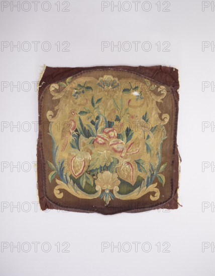 Tapestry upholstery for the seat and backrest of an armchair, unknown author, unknown provenance; flowers, floral ornaments, shells, birds; interior furnishings, furniture upholstery, decorative fabric tapestry; a seat, b backrest. Surrounded by a frame with motifs of acanthus leaves, shells, and flowers. Colors: brown and yellow background, red, dirty pink, sky blue, green, and yellow-green ornament. Late 18th century, France. Fabrics; height 59 cm, width 57 cm., A square textile with a worn, decorative woven panel featuring a central floral arrangement framed by a scrolling cartouche; the fabric shows frayed edges and a few small holes. The visible colors include shades of brown and tan in the background and border, muted gold and yellow tones in the central cartouche, and green and blue hues in the leaves and foliage. There are accents of red and pink in some of the flower forms and pale beige in other floral elements, along with small areas of darker brown and olive. The piece rest...