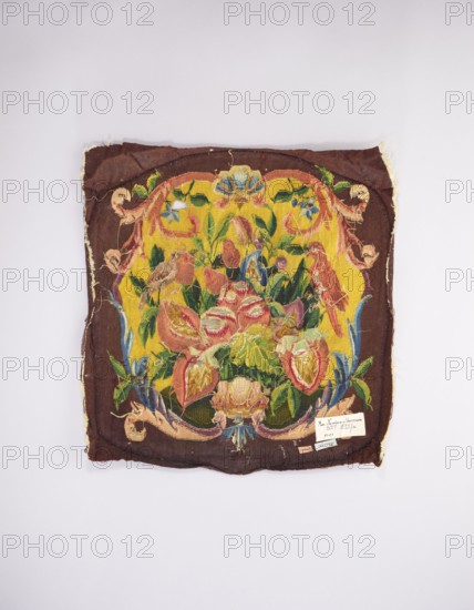 Tapestry upholstery for the seat and backrest of an armchair, and provenance; design with flowers, floral ornaments, shells, birds; interior furnishings, furniture upholstery, decorative fabric tapestry; a seat, b backrest. Surrounded by a frame with motifs of acanthus leaves, shells, and flowers. Colors: brown and yellow background, red, dirty pink, sky blue, green, and yellow-green. Late 18th century, France. Fabrics; height 59 cm, width 57 cm., A square textile with a dark brown border and a bright yellow central field displaying a floral motif, showing a group of stylized flowers and leaves in shades of pink, red, green, blue and cream, with additional accents of pale blue and orange; the fabric appears frayed at the edges and has loose threads and small areas of wear, with decorative scrolling elements in muted pink and cream along the border and a small rectangular patch attached near one corner; the background around the textile is light gray.