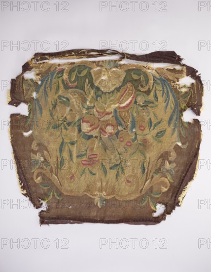 Tapestry upholstery for the seat and backrest of an armchair, and provenance; floral ornaments, shells, birds; interior furnishing upholstery, decorative fabric; a seat, b backrest. Surrounded by a frame with acanthus leaves, shells, and flowers. Colors: brown and yellow background, red, dirty pink, sky blue, green, and yellow-green. Late 18th century, France. Fabric; height 59 cm, width 57 cm., A worn rectangular textile with frayed edges and several holes lies flat against a pale background; its central area shows a faded decorative motif in tones of gold and tan with accents of green, blue, and muted red, surrounded by a darker brown border; the design includes leafy shapes, curved ornamental forms, and rounded fruit-like elements, with some areas where the weave is thin or missing and the underlying threads are exposed.