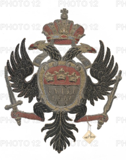 Coat of arms cartouche, restitution, embroidery: heraldic double-headed eagle bearing coats of arms; on its chest a shield with upper part red field bearing three crowns in a row, lower part silver field with white and red ermine tails. The eagle holds a sword in one talon and a scepter in the other. Embroidery colors: gold, silver, black, red, sky blue, and gold. Probably the coat of arms of Cologne. 18th century, Germany. Fabrics; height 26 cm, width 21 cm., An embroidered heraldic emblem depicting a double-headed eagle with outstretched wings, rendered mainly in black with gold outlining and feather details, a central shield area with red and gray cloth, gold ornamental embroidery around the shield and crown, and accents of red on beaks and ribbon-like elements; a gold and red crown sits above the eagle, and the eagle holds objects at each side with gray and gold tones.