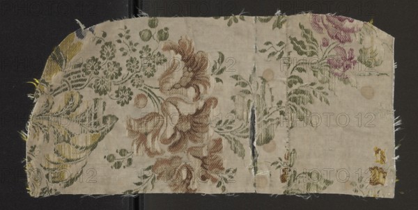 Fabric fragment, fabrics, fragments of silk fabrics, patterned fabrics, 18th-century silk fabrics, floral patterns. White grosgrain background, pattern displayed and brocaded with threads. Brocade threads: celadon, lemon yellow, ink, pink, orange, pale pink, in a faded red dry rose color tied with a twill weave. Brocade threads - green. Motifs: floral sprigs forming a kind of winding S-shaped tendril. Tiny flowers and fruit combined with sprigs of blooming, heavy flowers with calyxes resembling lilies or tulips. Expressive, full flowers with a soft outline. 18th century, France; height 39.9 cm, width 19 cm., A rectangular piece of fabric with frayed edges lies against a dark background, showing a floral woven pattern. The cloth ground is a light beige or off-white, with floral motifs in green, brown, muted pink, yellow, and a few darker olive tones. The left side shows a large leaf-like design in green and yellow, the central area has sweeping petal shapes in shades of brown and tan su...