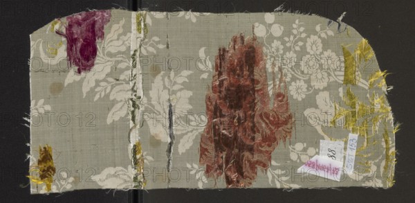 Fabric fragment, 18th-century silk fabric, France. White grosgrain background, pattern displayed and brocaded with threads. Brocade threads: celadon, lemon yellow, ink, pink, orange, pale pink, faded red dry rose color tied with a twill weave; brocade threads green. Motifs: floral sprigs forming winding S-shaped tendrils; tiny flowers and fruit combined with sprigs of blooming, heavy flowers with calyxes resembling lilies or tulips; expressive, full flowers with a soft outline. Height 39.9 cm, width 19 cm., A rectangular fabric fragment with frayed edges placed on a dark background, showing a pale gray-green base with an overall floral damask pattern in an off-white shade; several areas of shiny, textured velvet patches appear in deep burgundy, rust red, and golden yellow, with one irregular vertical strip of pale silver and green threads; a few small torn threads and worn spots reveal underlying weave, and a small folded paper object rests near one edge with visible color but no trans...
