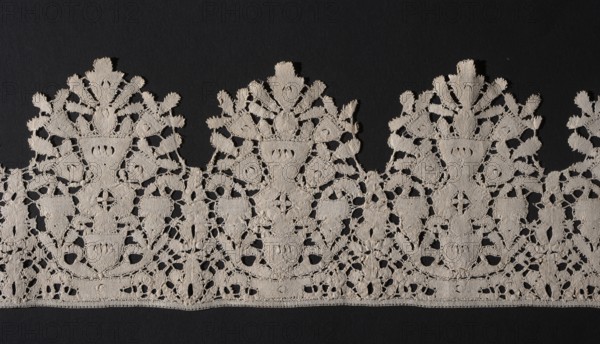 Lace fragment, Floral ornaments. Lace, lower edge serrated. In each oval there is a vase of flowers; flower crowns overlap the serrations. Joined with spiderwebs, so-called Van Dyck. 1630–1650, Flanders. Lace; length 200 cm, width 13.5 cm., A continuous band of ornamental lace in a light beige color is set against a dark black background; the lace displays a repeating scalloped top edge with ornate floral and vase-like motifs, openwork areas, and densely stitched sections creating textured patterns; the bottom edge is straight with a narrow finished hem, and the overall piece shows intricate looping and connecting threads with a combination of solid and airy elements in beige and black.