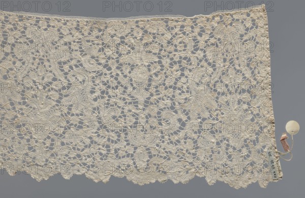 Lace fragment., With floral vase motifs with flower branches in a candelabra arrangement. Joints with picots. Gently serrated lower edge. Lower edge with a ladder. 17th century, Brussels. Materials: lace, fabric. Width 21 cm, length 162 cm., A rectangular piece of openwork lace with an intricate floral and scroll pattern in an off-white or cream color set against a gray background, showing a scalloped lower edge and a straight top edge; a small round off-white disk and a few attached small tags in beige and brown hang from the right edge.