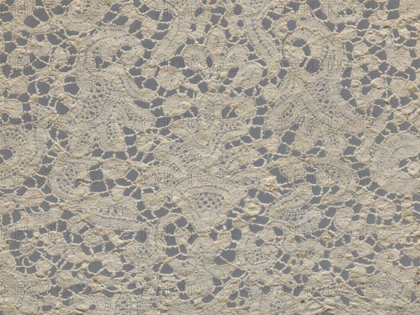 Lace fragment., With floral vase motifs with flower branches in a candelabra arrangement. Joints with picots. Gently serrated lower edge. Lower edge with a ladder. 17th century, Brussels. Lace, fabric; width 21 cm, length 162 cm., A close-up view of a piece of lace fabric showing a repeating openwork pattern with irregular holes and interconnected motifs, made of off-white or cream-colored threads against a muted gray background; the lace has a textured, slightly raised surface with areas of denser stitching and finer net-like sections, and there are a few small darker brownish spots scattered on the fabric.