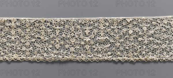 Lace fragment, lace with a fine floral pattern; de brides background with picots; lower edge with fine serrations, upper edge straight; 17th–18th century, Venice; materials: lace fabric; dimensions: width 9.5 cm, length 134 cm., A horizontal strip of delicate lace laid flat against a smooth surface, showing an openwork pattern of interconnected loops and floral motifs with a scalloped edge; the lace is a creamy off-white color and the background surface is a medium gray, creating contrast between the intricate textile and the flat backdrop.