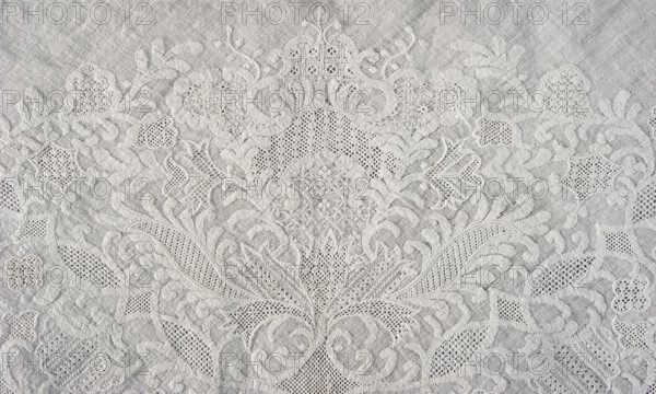 Shawl, fashion accessory, clothing, women's clothing, 1st half of the 19th century; 18th century fashion accessories, white embroidery, Dresden lace, plant and floral patterns, Ornament composed symmetrically around an axis passing through the centre of the shawl, occupying almost the entire surface. On the edge of two hypotenuse sides a geometric ornament: knitted serrations on the edge, above it a strip of fine mesh, separated from the plant ornament filling the space of the shawl by a series of arched forms. Silesia place of origin, Saxony place of creation, height 79 cm, width 34 cm, The image shows a textured fabric with an ornate raised pattern featuring floral and leafy motifs and areas of openwork. The design forms a symmetrical, intricate arrangement with curved lines, scalloped edges, and small perforated sections. The visible colors are shades of white and light gray, creating subtle contrast between the raised embroidery and the background cloth.
