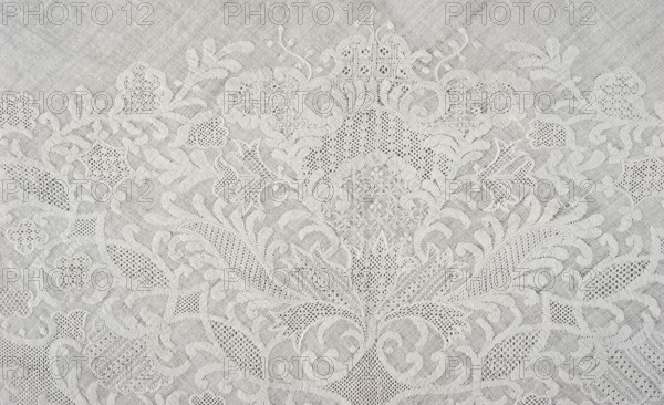 Shawl, repossession provenance; shawl; fashion accessory; clothing; women's clothing; 1st half of the 19th century; 18th century; white embroidery; Dresden lace; plant and floral patterns; Saxony Germany; Silesia place of origin; ornament composed symmetrically around an axis passing through the centre of the shawl occupying almost the entire surface; on the edge of two hypotenuse sides a geometric ornament: knitted serrations on the edge, above it a strip of fine mesh, separated from the plant ornament by a series of arched forms; height 79 cm, width 34 cm., A close-up view of a textured fabric with an intricate floral lace pattern, showing a group of stylized flowers, leaves, and curling vine-like shapes in a repeating ornamental design, rendered in shades of white and off-white with subtle shadows that create depth and highlight the raised and openwork areas.