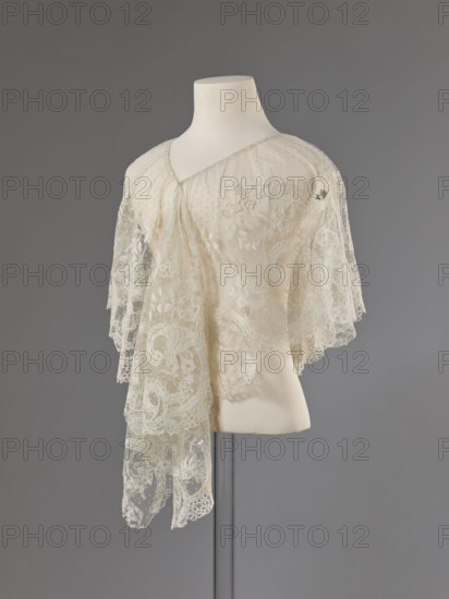Collar, Lace collar so-called berta with a pattern of flowers, leaves and small dots in the background; floral and plant ornaments; clothing accessory for women's fashion; lace; width 33 cm; dated 1840–1860; possibly Brussels;, A mannequin torso draped with a delicate lace garment featuring sheer layers and intricate floral and paisley-like patterns, the lace appearing in shades of ivory and cream with translucent areas and fine dotted mesh, the garment forming soft cascading edges and scalloped hems, displayed against a smooth neutral gray background with the mannequin mount and a slender support pole partially visible in light off-white and muted gray.