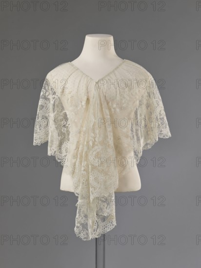Collar, lace collar so-called berta with a pattern of flowers, leaves and small dots in the background; floral and plant ornaments; clothing accessory for women's fashion; 1840–1860; Brussels, lace; width 33 cm., A mannequin torso displays a delicate lace shawl or capelet in shades of cream and off-white draped over its shoulders, with sheer floral lace panels and scalloped edges that hang down in a layered, flowing arrangement; the mannequin is white and mounted on a thin stand against a smooth gray background.