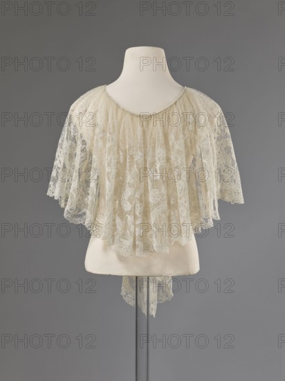 Collar, Lace collar so-called berta with a pattern of flowers, leaves and small dots in the background; floral and plant ornaments; clothing accessory for women's fashion; lace; width 33 cm; dated 1840–1860; probably Brussels;, A mannequin torso in off-white displays a delicate capelet made of sheer lace in pale cream and ivory tones, with a scalloped lace edge and floral lace patterns throughout; the lace is gathered at the neckline and falls in soft pleats over the shoulders and upper back, creating a layered, translucent effect against a plain gray background, with the mannequin mounted on a slender dark pole and a small piece of lace hanging down beneath the hem.