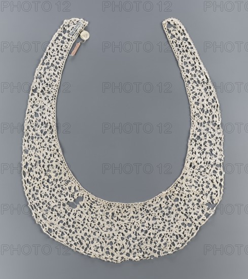 Collar, lace, Kublicki Piottuch, Alfred 1861-1945 — lace collar, clothing accessory. The ribbon pattern imitates a twig or floral sprig. Oval collar with very elongated ends. 17th century, Italy, lace, clothing accessory, width 11 cm, A delicate crocheted collar in an off-white or cream color is displayed against a smooth medium-gray background; the collar has a wide crescent shape with a fine openwork pattern of interconnected loops and floral-like motifs, tapering into two narrower curved ends, and a small round pale-colored tag and a tiny rectangular tag are attached near one end.